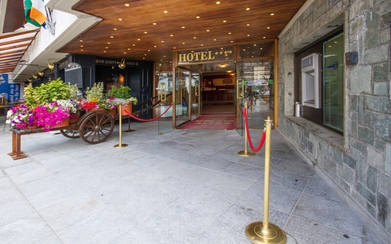 Hotel entrance with stone exterior, wooden ceiling, gold "HOTEL" sign, red carpet, and potted plants under a canopy.