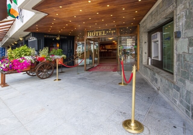 Ingresso di un hotel con insegna "HOTEL", porte a doppio battente, tappeto rosso e carretto fiorito. Atmosfera accogliente.