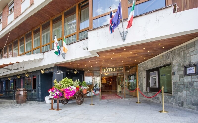 Exterior of a hotel with a glass door, red carpet, flags, and flower cart, showcasing an elegant and inviting atmosphere.