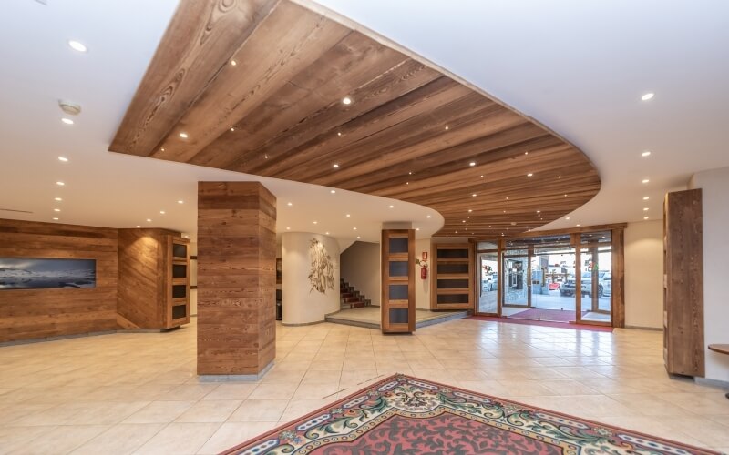Spacious room with high ceilings, light tiles, red rug, wooden pillars, TV, staircase, and glass doors leading outside.