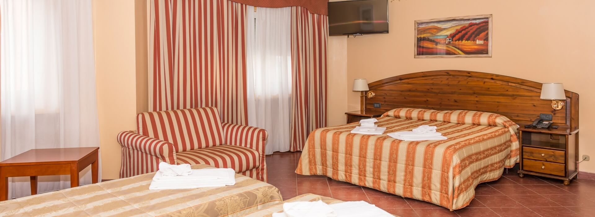 Cozy hotel room with two beds, a chair, table, peach walls, red tile floor, and warm lighting from bedside lamps.