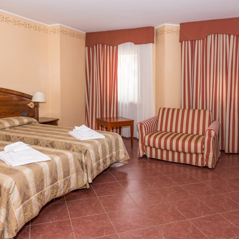 Camera d'hotel con due letti, poltrona a righe, tavolo, decorata in rosso e beige, con pavimento in piastrelle rosse.