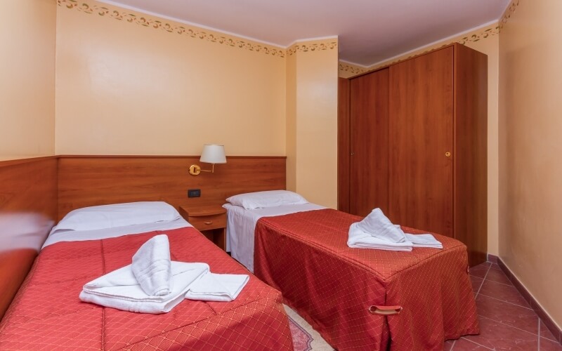 Cozy room with two beds, red blankets, white towels, wooden headboards, and a small table with a lamp between them.