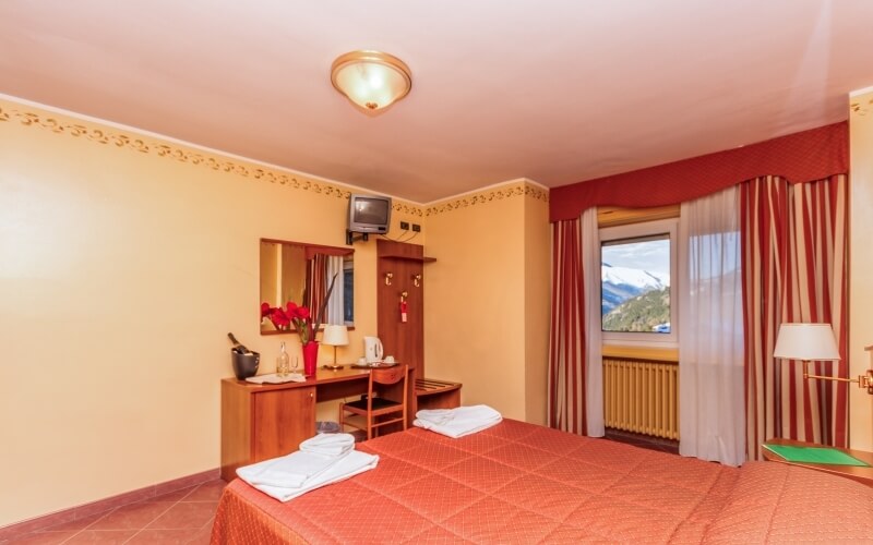 Cozy bedroom with a red blanket on the bed, dresser with a mirror, and a window showing a mountainous landscape.