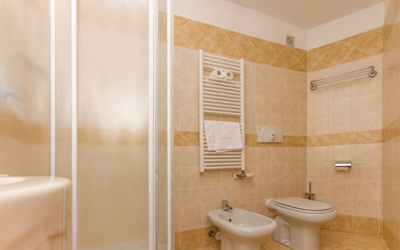A clean bathroom featuring a glass shower, white toilet, bidet, radiator, and brown-tiled walls and floor.