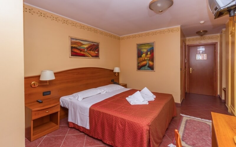 Cozy hotel room with a bed, nightstands, dresser, and artwork, featuring a red quilt and swan towels on the bed.
