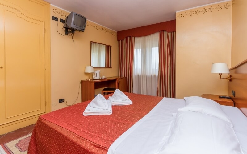 Cozy hotel room with a large bed, yellow wardrobe, wooden tables, TV, kettle, and striped curtains.