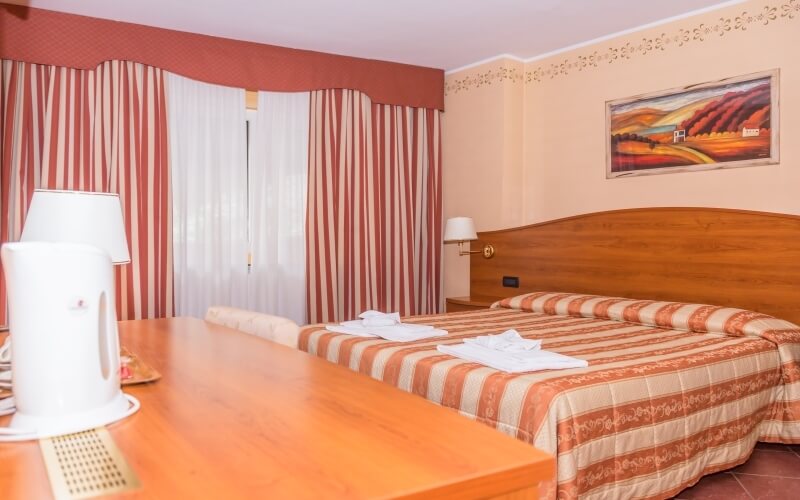 Cozy hotel room with two beds, striped bedding, a table, lamp, and a landscape painting above the headboard.