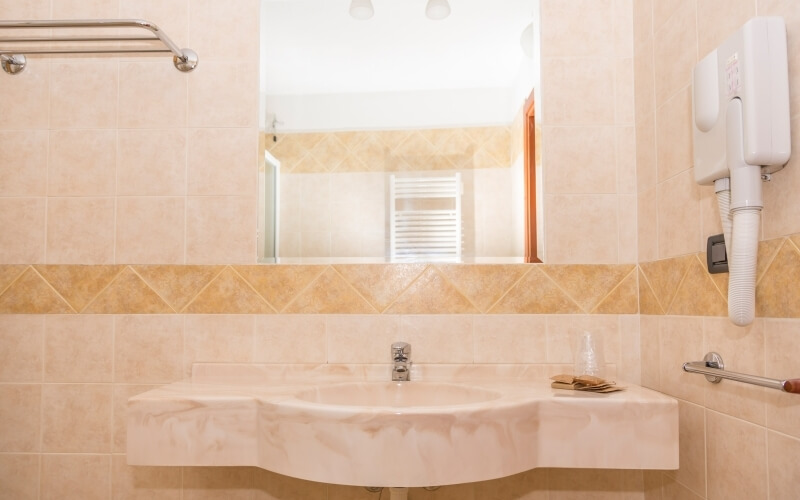 Bagno con piastrelle beige, lavabo in marmo bianco, specchio grande, asciugamani e asciugacapelli a parete.