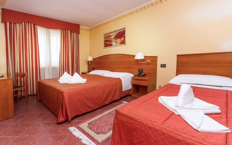 Cozy hotel room with two beds, red blankets, a window with striped curtains, and a wooden desk, featuring a warm color scheme.