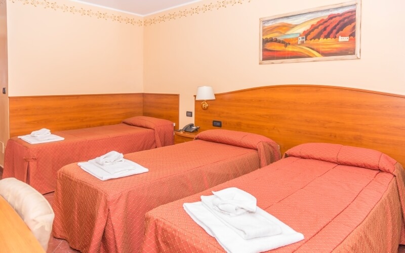 Hotel room with three beds, red bedspreads, white towels, wood panel wall, lamp, telephone, and beige walls.