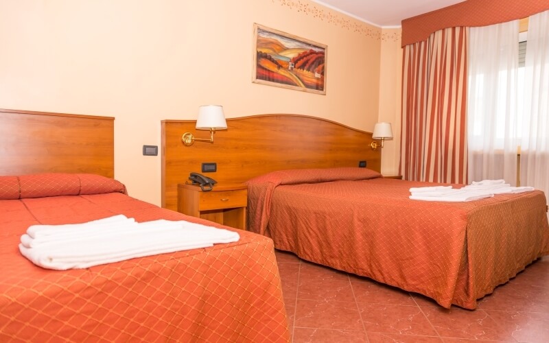 Cozy hotel room with two beds, red bedspreads, lamps, a nightstand, and a window with sheer curtains and drapes.