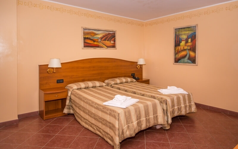 Cozy hotel room with two beds, striped comforters, nightstands, lamps, paintings, warm peach walls, and brown tiled floor.