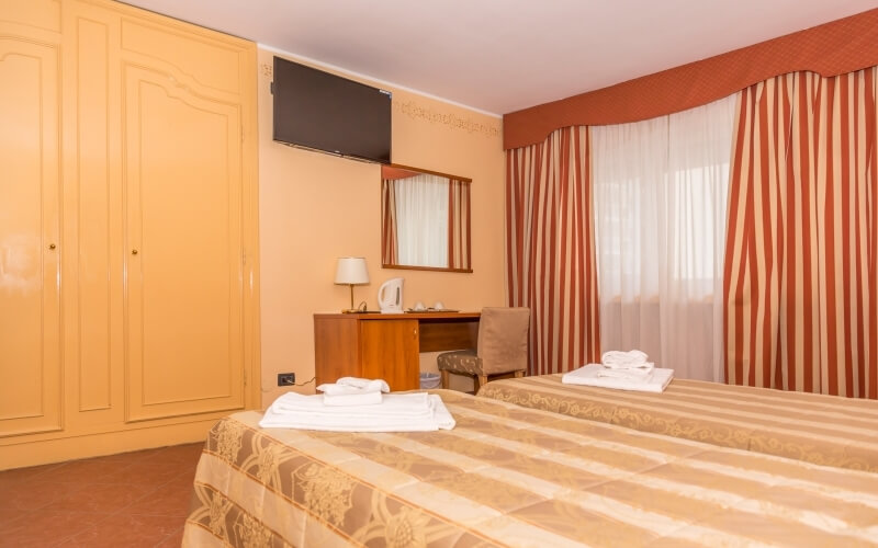 Hotel room with two floral beds, a desk, mirror, TV, wardrobe, and peach walls, exuding cleanliness and organization.