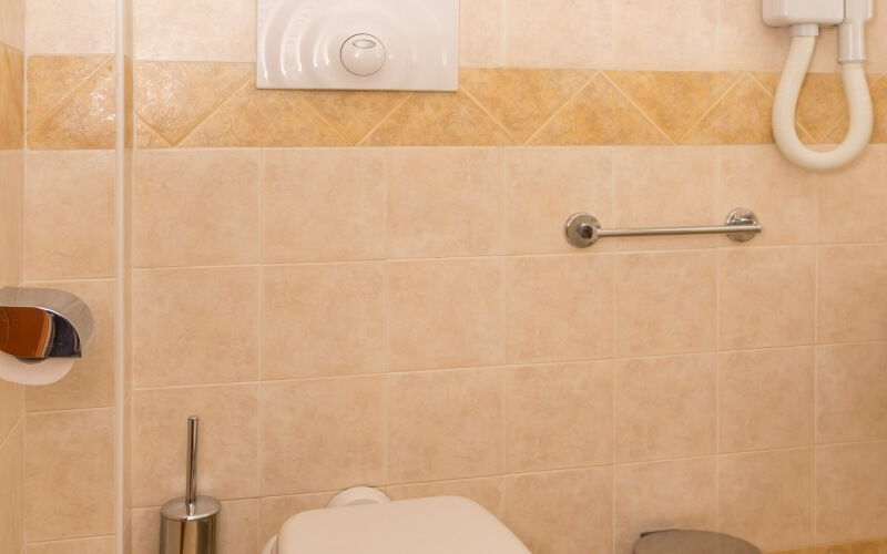 Bathroom scene featuring a white toilet, light-brown wall tiles, grab bar, bidet attachment, toilet paper holder, and trash can.