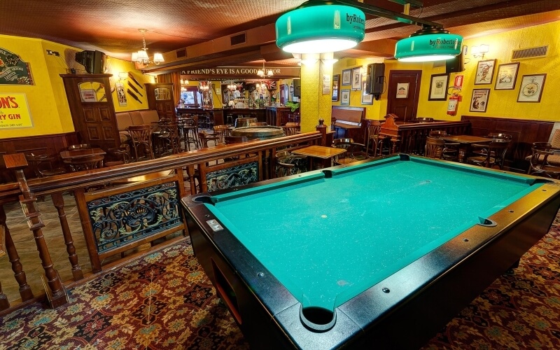 Un tavolo da biliardo verde illuminato da lampade turchesi in un bar con pareti gialle e decorazioni varie.