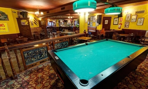 Bar con tavolo da biliardo, atmosfera accogliente, luci arancioni, schermo per partite di calcio e decorazioni eclettiche.