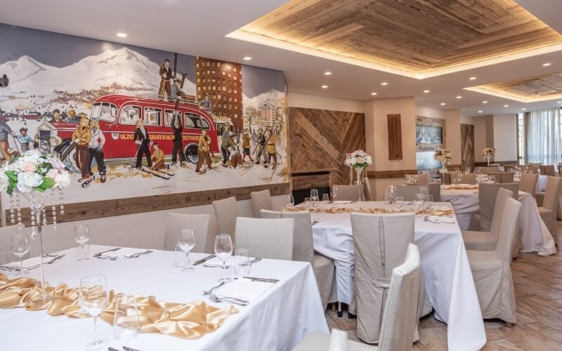Formal restaurant setting with white tablecloths, a mural of a red bus and mountains, and elegant table settings.
