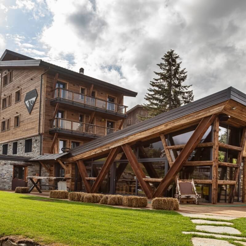 Large wooden building with a sloping roof, balcony, manicured lawn, hay bales, and patio chairs under a cloudy sky.