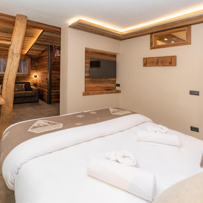 Serene bedroom with beige walls, a bed with rolled towels, a mounted TV, and a doorway leading to another room.