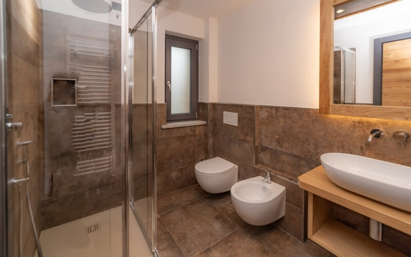 Bagno moderno con doccia in vetro, WC, bidet, lavabo e specchio, pavimento in piastrelle marroni e pareti bianche.