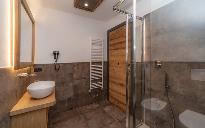 Bagno moderno con doccia in vetro, lavabo bianco, specchio elegante e pareti in piastrelle marroni e bianche.