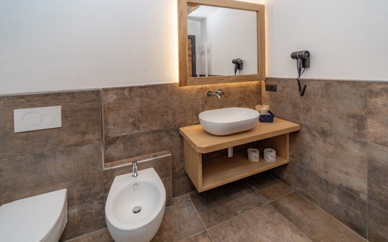 Bagno moderno con bidet bianco, lavabo su piano in legno, specchio illuminato e asciugacapelli nero attaccato al muro.