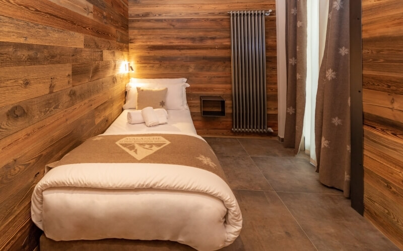 Camera accogliente con letto angolato, pareti in legno chiaro, pavimento in piastrelle scure e finestra con tende a fiocchi di neve.