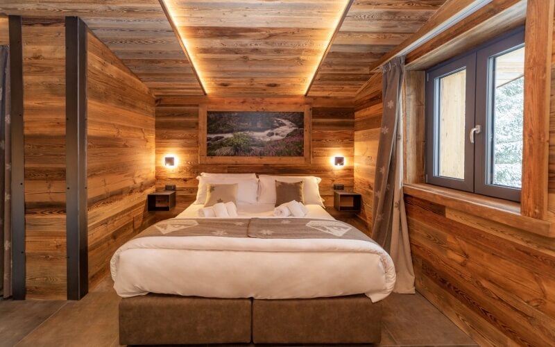 Cozy bedroom with a large bed, wooden walls, nightstands with lamps, brown ottoman, and a window with outdoor views.