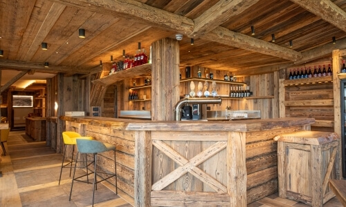 Area bar rustica con un bancone in legno, scaffali di bottiglie e un'atmosfera accogliente e illuminata.