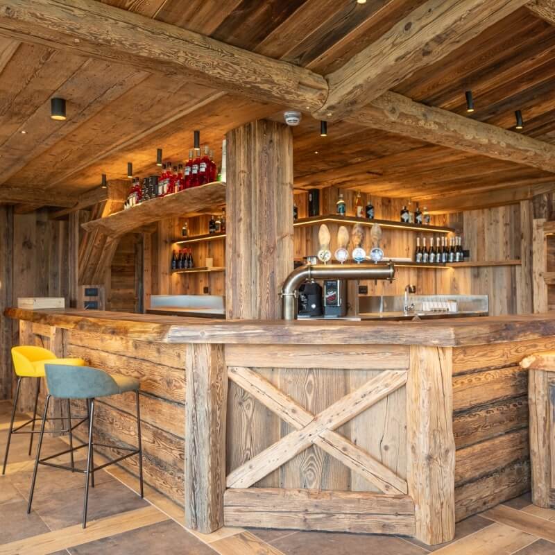 Rustic bar with wooden countertop, X-shaped front, yellow and blue-gray chairs, and shelves of assorted liquor bottles.