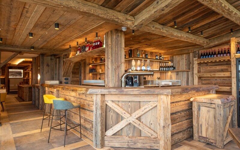 Rustic wooden bar with two barstools, shelves of liquor, and artwork, illuminated by black lights and a surveillance camera.