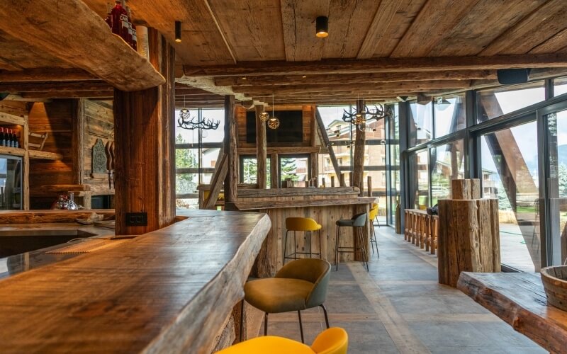 Bar rustico in legno con sedie gialle, lampadario di corna, pareti in legno e una grande finestra che offre vista panoramica.