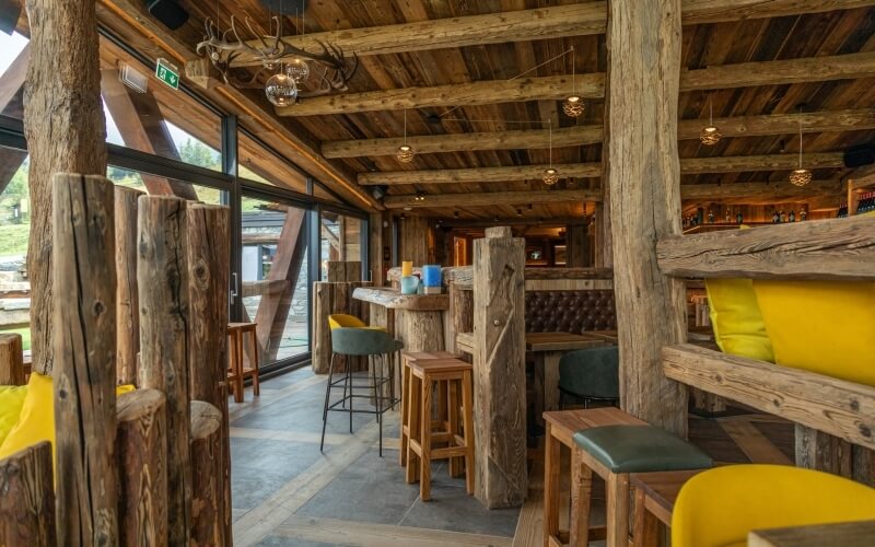 Rustic dining area with wooden beams, large windows, mixed seating, and antler chandeliers, creating a cozy ambiance.