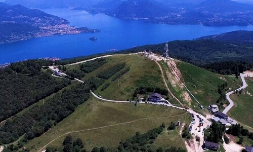 Aerial view of a grassy hill with a metal tower, buildings, roads, trees, and a lake in the background.