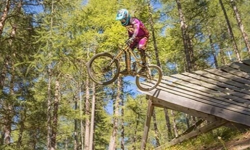 Mountain biker mid-air on a wooden ramp in a forest, wearing a blue helmet, pink shirt, and red shorts, with sunlight filtering through trees.