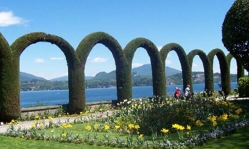 Sunlit garden archway with shrubs and flowers, leading to a blue body of water and hilly green landscape under a clear sky.