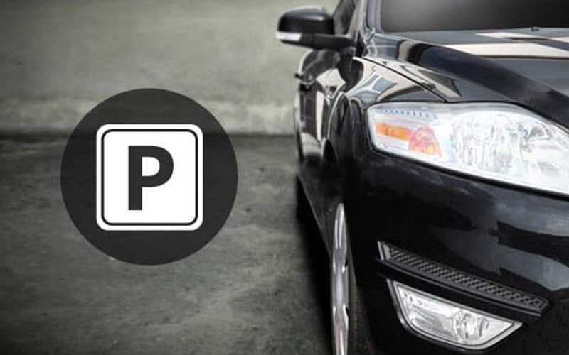 Parking sign with a "P" next to the front end of a shiny black car in a grey cement parking lot.