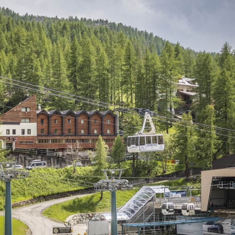 Sistema di funivia in montagna con hotel "HOTEL MONTANA" e altri edifici, circondato da alberi e verde.
