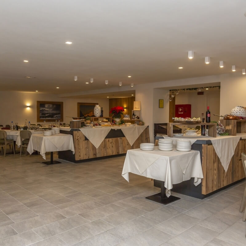 Ristorante con area buffet ben illuminata, tavoli con tovaglie bianche e un buffet di cibo su tavolo in legno.