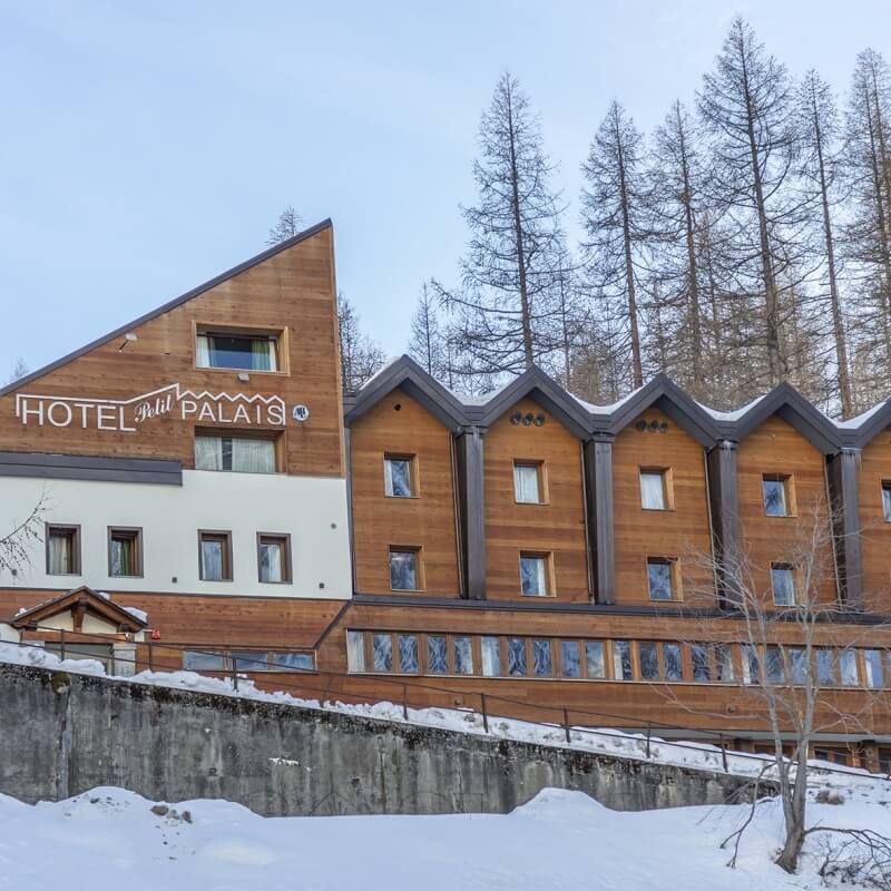 Exterior view of Hotel Petit Palais with wooden and white design, surrounded by snow, trees, and an overcast sky.