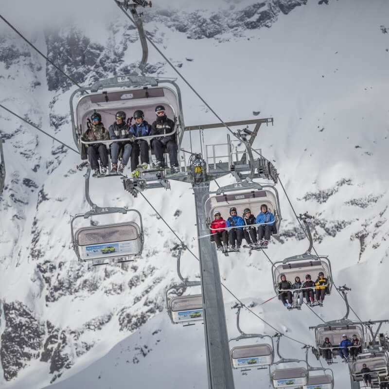 Immagine di una seggiovia con sciatori su una montagna innevata, circondata da alberi e rocce. Atmosfera invernale.