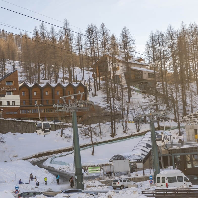 Paesaggio montano innevato con un impianto di risalita, edifici e alberi spogli sotto un cielo sereno.