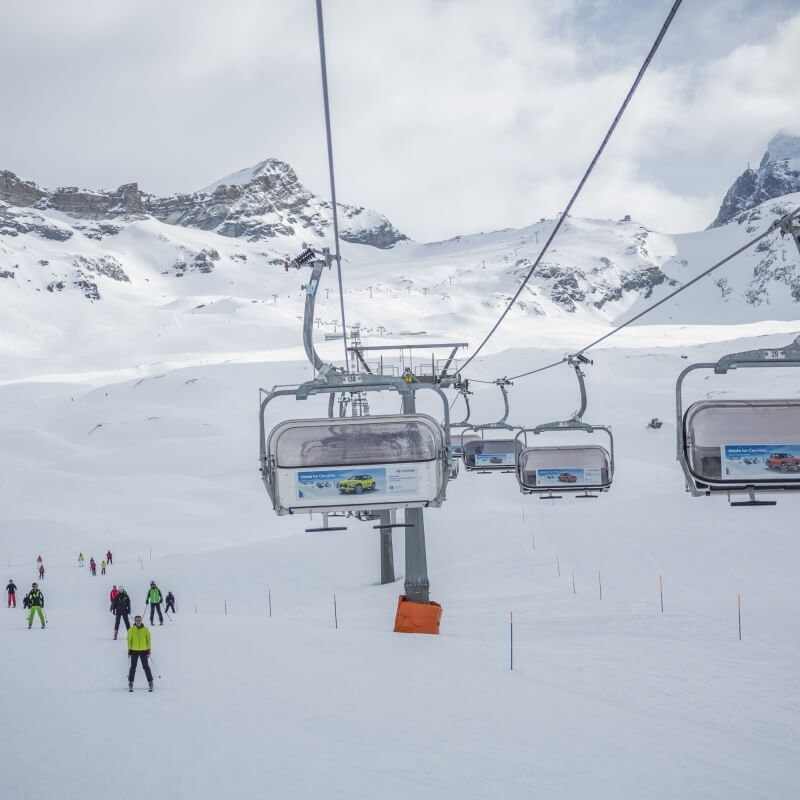 Pista da sci innevata con seggiovia e sciatori, montagne rocciose sullo sfondo e cielo nuvoloso.