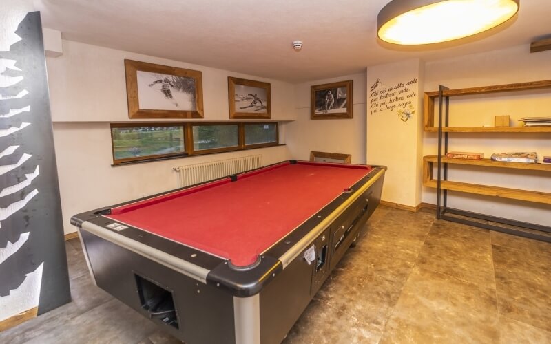 A game room featuring a black pool table with red felt, white walls, artwork, a bookshelf, and warm lighting.