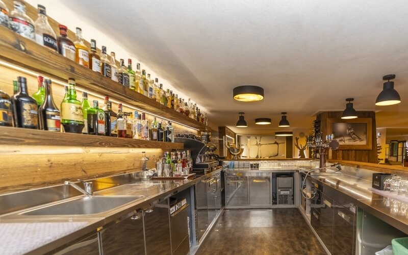 View from behind a stainless steel bar counter with bottles, a double sink, and a glossy white bar top with mounted horns.