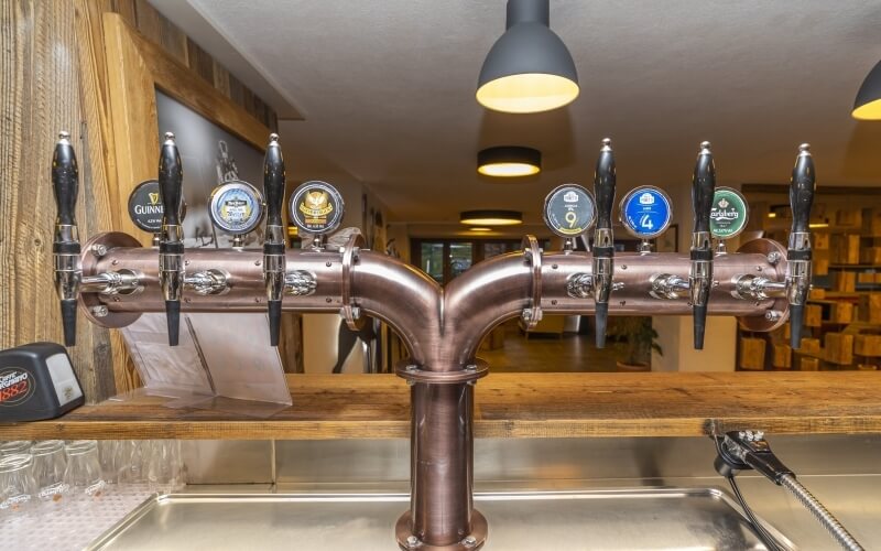 Bar with copper beer taps, eight brands displayed, stainless steel sink, wooden wall sign "Fischer's," and a hallway in background.