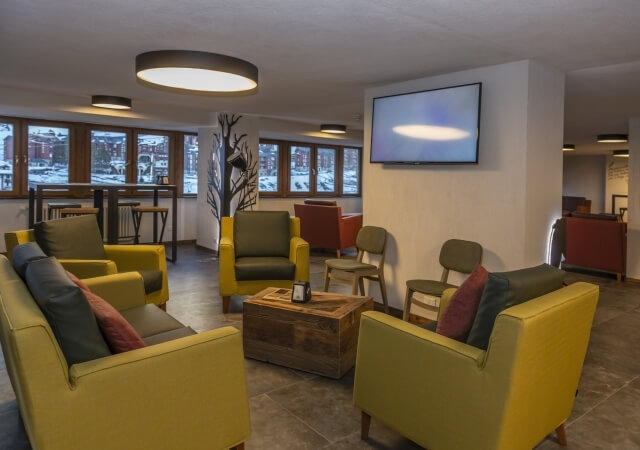 Modern minimalist lobby with yellow armchairs, wooden tables, large TV, and snow-covered view through windows.