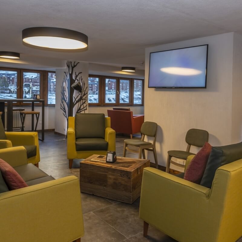 Modern room with yellow seating, large windows, a TV, and a blend of natural and industrial decor, overlooking snowy outdoors.