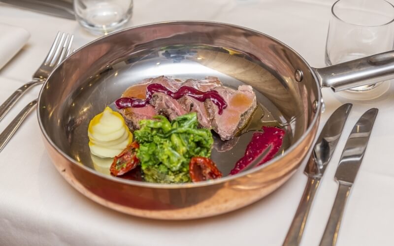 Table setting with a stainless steel pan of sliced meat, broccoli, and potatoes, accompanied by silverware and glasses.